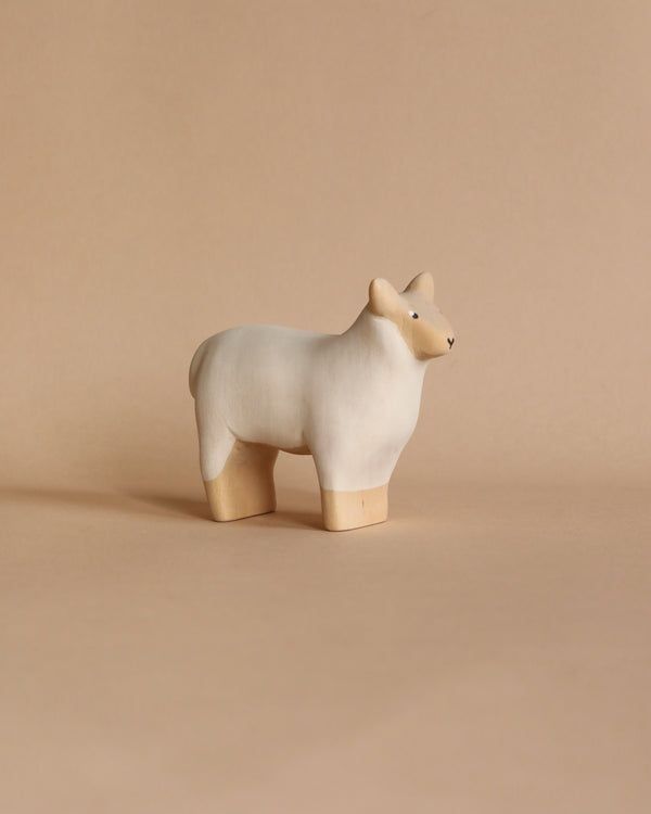 A small, minimalist ceramic sculpture resembling a handmade wooden sheep, with a light beige body and slightly darker legs and face, placed on a matching beige background.