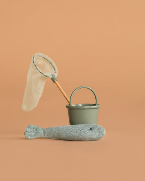 The Maileg Fishing Set, Mouse features a small green bucket, a toy fishing net with a wooden handle, and a soft stuffed fish—perfect play accessories for mouse families—arranged on a plain beige background.