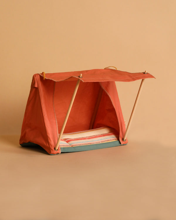 The Maileg Happy Camper Tent - Rust is a compact, foldable tent with vibrant orange fabric and wooden poles. It includes a red and white striped mat, set against a plain beige background, making it resemble a cozy miniature beach cabana ideal for indoor sleepovers.