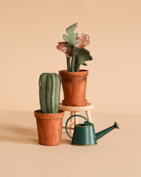 The Maileg Gardening Set, featuring three small potted fabric plants in miniature pots and a green watering can, is displayed on a light beige surface with one plant elevated like mouse furniture against a plain beige background.