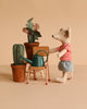 A Maileg Little Green Thumb Garden Set features a mouse in a red striped shirt and blue shorts standing by a flower pot and a green watering can on a school chair, all set against a beige background.