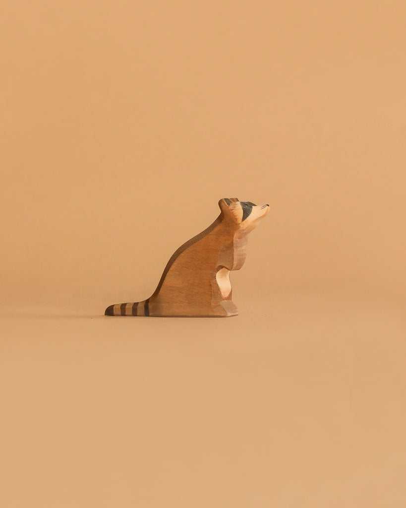 A handcrafted raccoon figurine from the Ostheimer Handmade Wooden Woodland Set sits on a light brown surface with a beige background, featuring hand-painted face and tail details and facing right.