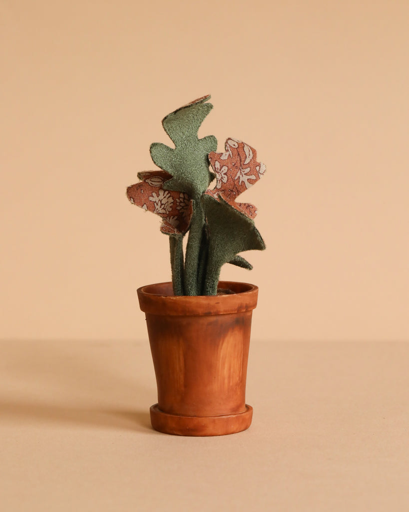 The Maileg Flower Pot With Plant, Miniature features a small brown pot with a fabric plant showcasing green and patterned leaves, set against a plain beige background for an authentic look.