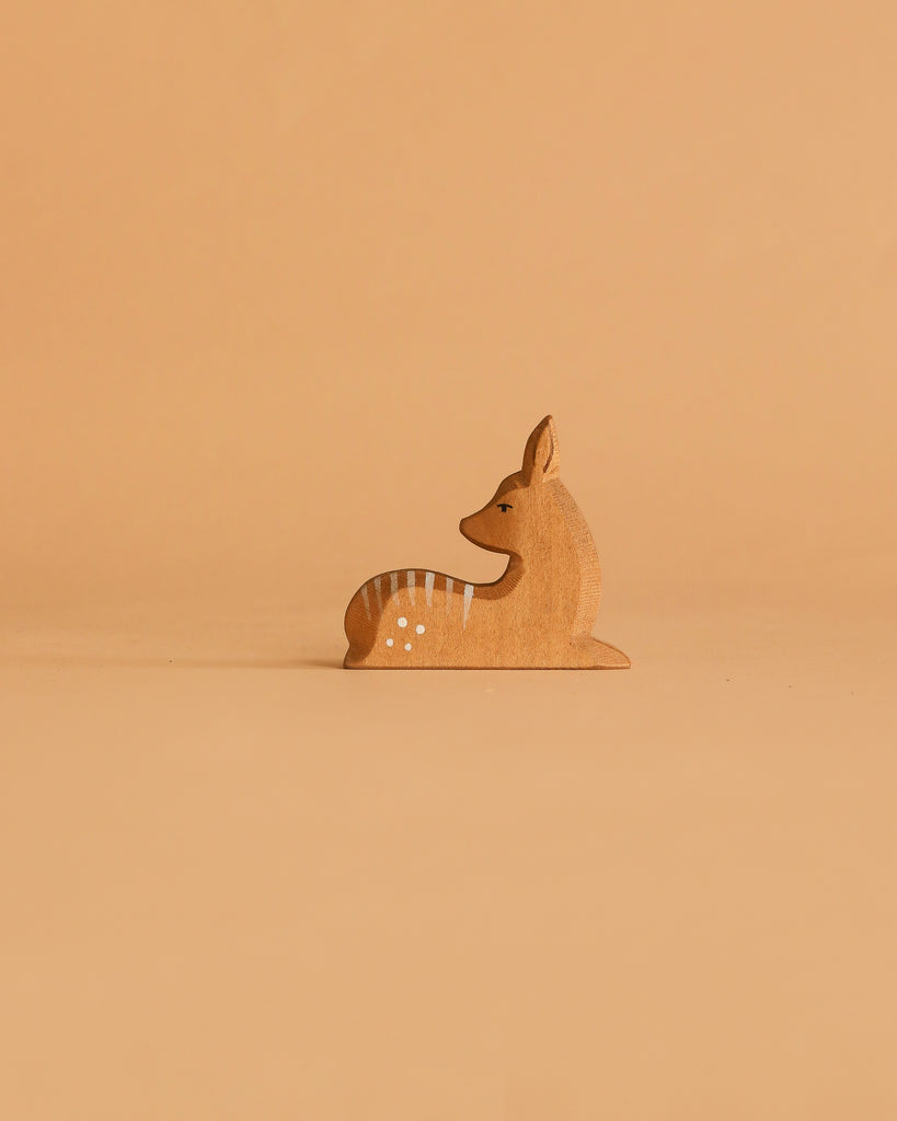 A small, handcrafted deer figurine with white spots from the Ostheimer Handmade Wooden Woodland Set sits against a light brown background, showcasing the timeless charm of these classic toys.