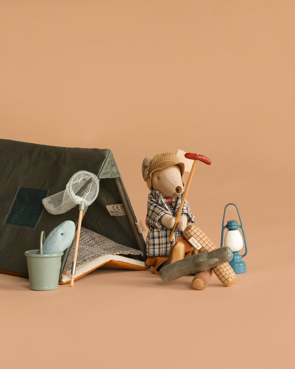 The Maileg Camping Set with Granddad features a toy mouse in a plaid coat by a cozy tent, holding a red-handled tool. Classic accessories—like a butterfly net, bucket with toy fish, blue lantern, and campfire—complete the beige background scene.