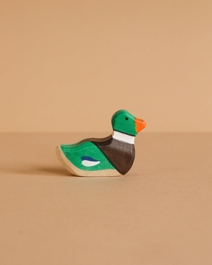 A small, handcrafted Holztiger Mallard Duck, Sitting—painted green, brown, white, orange, and blue—rests on a beige surface with a matching background, showcasing the charm and play value of HOLZTIGER figures.