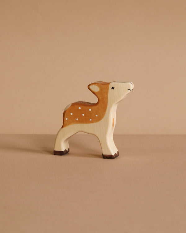 The Holztiger Fawn, a small handcrafted wooden deer with brown and white painted details, stands on a beige surface against a matching beige background.