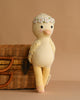 Cuddle + Kind Flora the Duckling - Ivory, a hand-knit duck with floral crown, leans on a wicker basket against a beige background—perfect for anyone seeking heirloom quality dolls.
