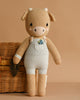 Cuddle + Kind Asher The Cow is a hand-knit light brown and cream cotton plush doll in white overalls, filled with hypoallergenic polyfill. It stands upright beside a woven wicker basket against a beige background.