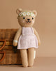 Cuddle + Kind Olivia The Honey Bear, a hand-knit teddy in a white dress and daisy flower crown, stands before a wicker basket against a beige background.