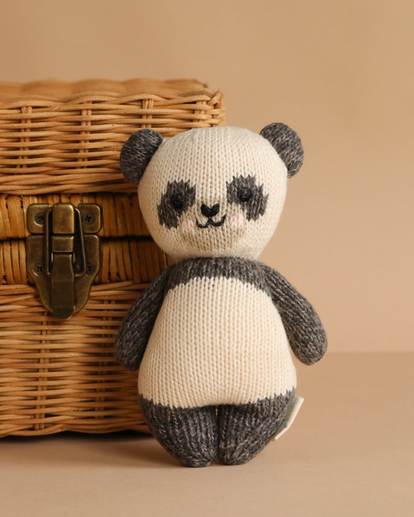 The Cuddle + Kind Baby Panda, an heirloom-quality hand-knit plush with dark gray ears, arms, and legs, is propped against a wicker basket with a brass latch on a beige surface and background.