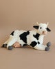 A Cow Stuffed Animal, featuring white and black spots, is lying down against a plain beige background. This artisan hand-sewn plush toy from our collection has small yellow horns, a pink udder, and grey hooves. Its ears and tail are black, matching some of the spots on its body.