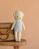 Cuddle + Kind Benjamin the Lamb, a hand-knit doll made from 100% cotton yarn with curly white wool and a blue and cream outfit, is shown leaning against a wicker basket on a wooden stool with a peach background.