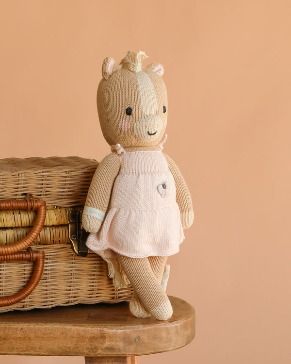 Cuddle + Kind Kelsie the Pony, a hand knit doll in a pale pink dress, leans against a wicker basket on a wooden stool with a soft peach backdrop. This fair trade treasure is lovingly handmade in Peru.