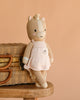 Cuddle + Kind Kelsie the Pony, a hand knit doll in a pale pink dress, leans against a wicker basket on a wooden stool with a soft peach backdrop. This fair trade treasure is lovingly handmade in Peru.