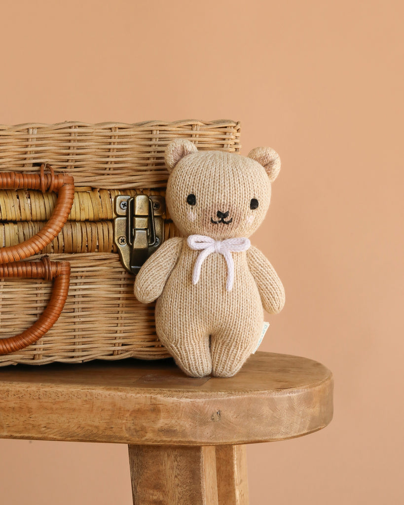 The Cuddle + Kind Baby Honey Bear, an heirloom-quality doll with a lavender bow, sits on a wooden stool beside a wicker basket against a simple beige background.