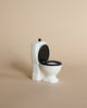 The Maileg Big Sister Bathroom Set features a small white toy toilet with a black seat and lid, displayed on a plain beige surface—perfect as an accessory for your Maileg collection.