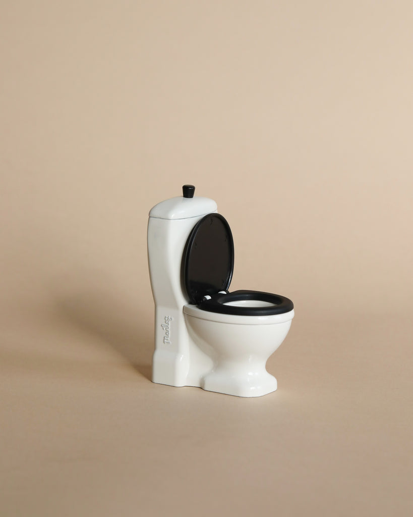 The Maileg Bathroom Set is a small white toilet with a black seat and lid, displayed on a beige surface—ideal for pairing with other Maileg mouse furniture like the bathtub or sink dresser.