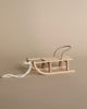 A small, wooden Maileg Christmas Mini Sled with a metal handlebar and a white rope attached to the front is placed on a neutral, beige background. This winter accessory features a simple, rustic design.