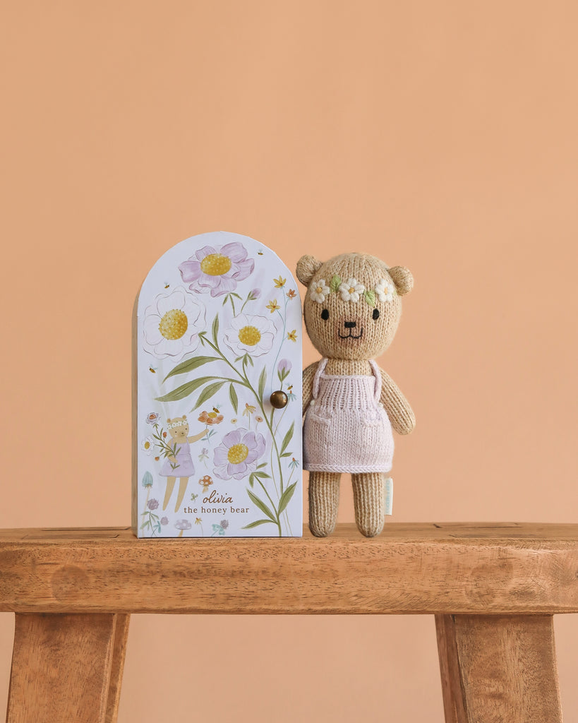 The Cuddle + Kind Tiny Olivia the Honey Bear, dressed in a light pink outfit, stands beside its floral illustrated box on a wooden bench set against a peach background.