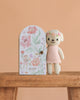 The Cuddle + Kind Tiny Charlotte The Dog, a hand-knit doll in a pink dress, stands beside a floral-patterned door labeled "charlotte the dog" on wood with a peach background. Handmade in Peru, this non-toxic toy is both charming and safe.