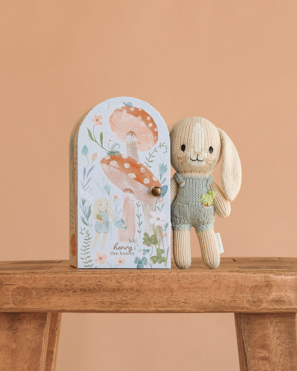 The Cuddle + Kind Tiny Henry The Bunny, a soft hand-knit toy in light beige and green, stands beside a pastel box with floral and mushroom designs on a wooden surface set against a peach background.