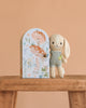 The Cuddle + Kind Tiny Henry The Bunny, a soft hand-knit toy in light beige and green, stands beside a pastel box with floral and mushroom designs on a wooden surface set against a peach background.