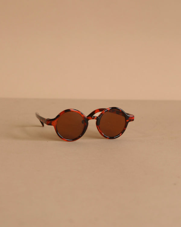 Minikane Doll Accessories | Harry Sunglasses feature round brown lenses and tortoiseshell frames, displayed on a beige surface—perfect stylish accessories for your Minikane doll.