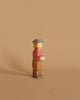 An Ostheimer Son, Gray Hat wooden toy figure with simplistic features stands against a beige background. It has blonde hair, a light gray hat, a red shirt, light brown pants, and brown shoes. Perfect for imaginative play, the handcrafted figure has a neutral expression and faces slightly to the right.