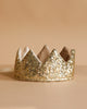 The Crown - Gold Sequins from Luciole et Petit Pois, inspired by France, features a pointed design with soft lining and sparkles brightly on a beige surface against a matching beige background.