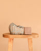 The Maileg Carry Cot, Large - Blue, featuring a quilted floral pattern and knitted strap, sits atop a wooden stool against a plain beige backdrop.