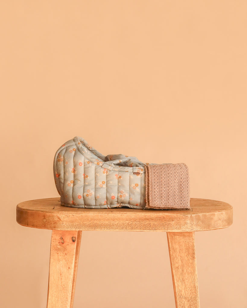 The Maileg Carry Cot, Large - Blue, featuring a quilted floral pattern and knitted strap, sits atop a wooden stool against a plain beige backdrop.