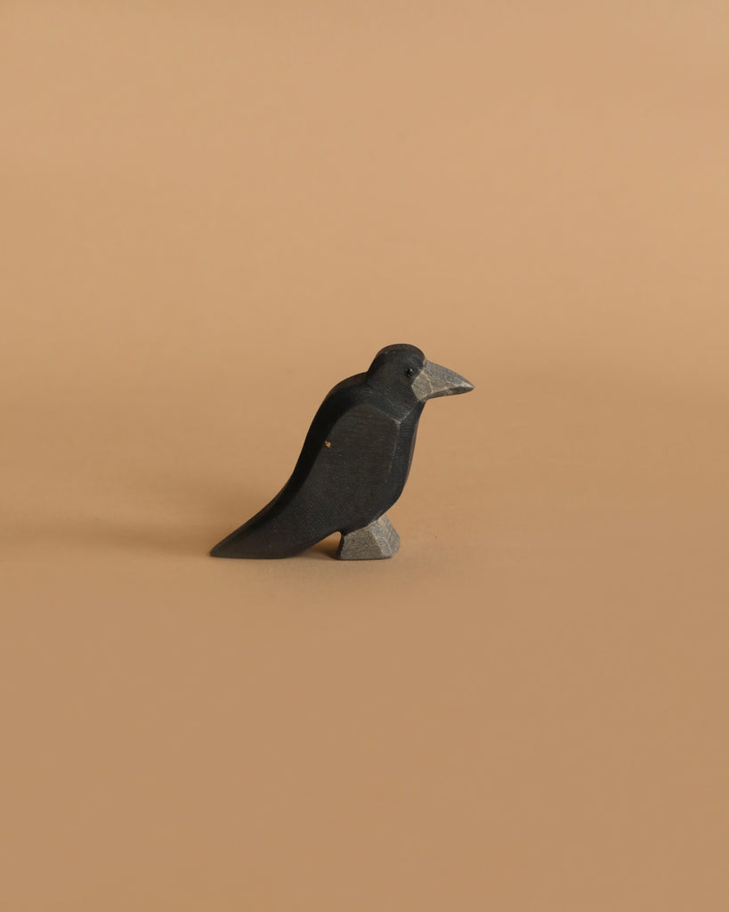 A small black bird figurine with a gray beak and gray legs is standing on a plain, light brown surface. The background is light brown, creating a minimalist and monochromatic setting. This Ostheimer Witch Set toy, crafted with sustainably sourced materials, embodies simplicity and elegance.