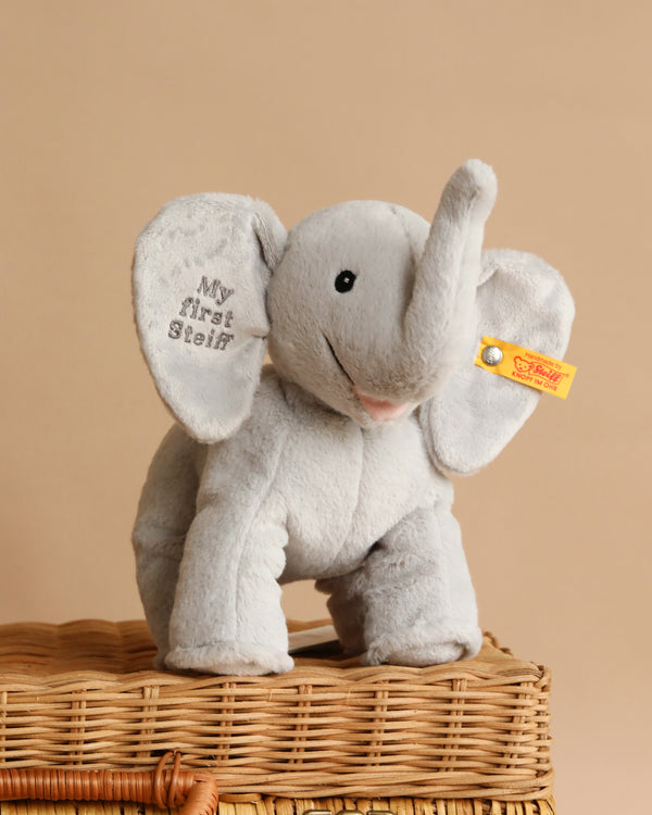The “My First Steiff” Ellie Elephant Plush Baby Toy (9 inches) is a soft, light gray elephant with "My First Steiff" embroidered on one ear and a yellow tag on the other—an adorable baby gift, shown sitting on a woven basket against a beige background.