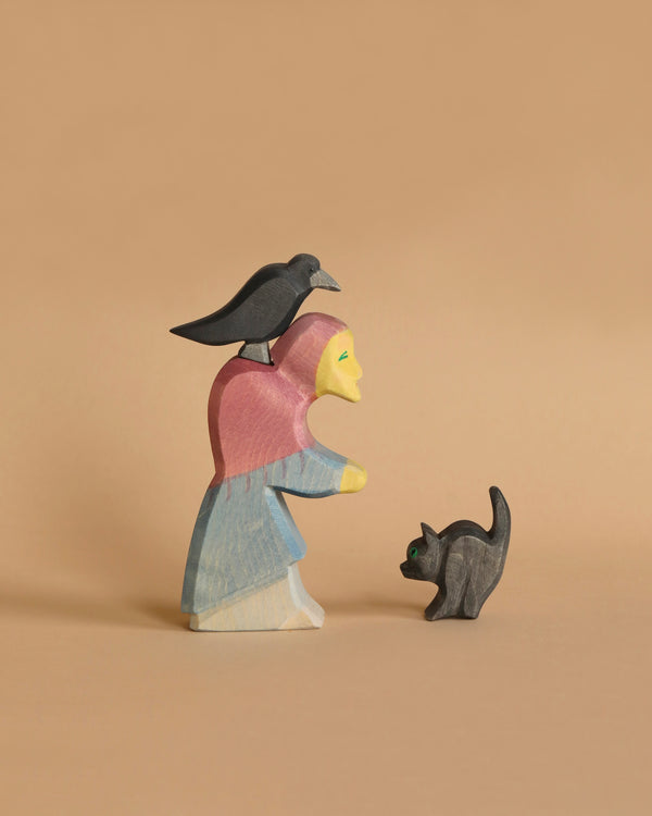 The Ostheimer Witch Set features a handcrafted wooden figure of a person wearing a pink headscarf and blue dress, made from sustainably sourced materials, standing on a beige background. A black bird perches atop the figure's head, while a small black cat with green eyes stands in front of them, gazing upward.
