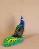 A colorful, whimsical Peacock Stuffed Animal with a vibrant blue and green body and an embellished tail featuring eye-like patterns, positioned against a soft peach background.