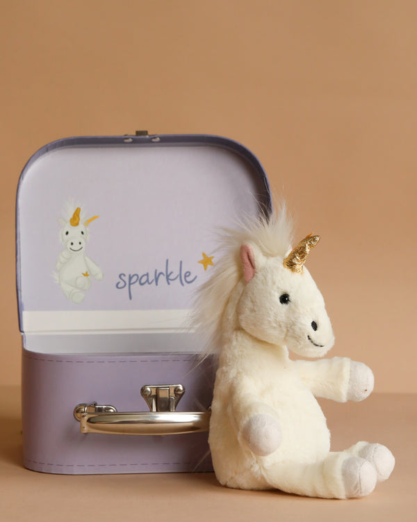 The Steiff Unica Dangling Plush Toy Unicorn in Suitcase features a plush unicorn with a golden horn, seated before its open case. Inside, a unicorn illustration and the word "sparkle" inspire magical adventures for little dreamers.