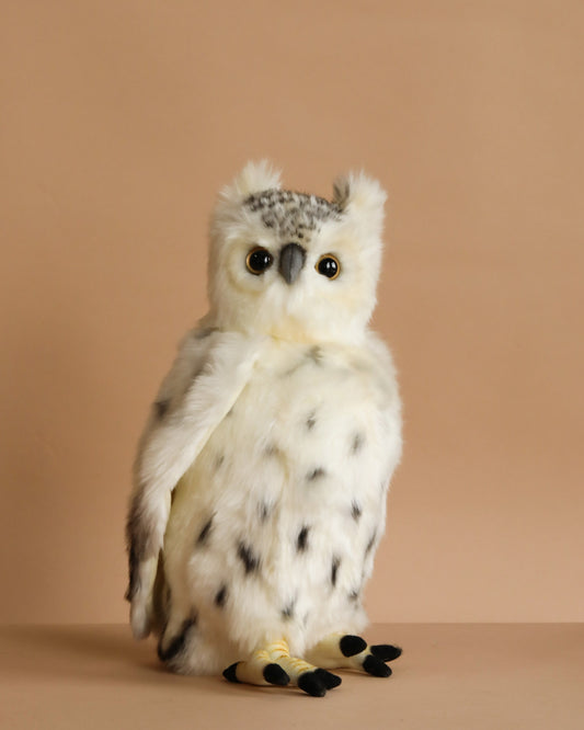 The Snowy Owl Stuffed Animal features hand sewn white and gray fur with black spots, round yellow eyes, and a small beak. It sits upright against a plain beige background, capturing the lifelike detail typical of HANSA creations.