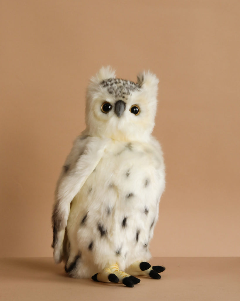 The Snowy Owl Stuffed Animal features hand sewn white and gray fur with black spots, round yellow eyes, and a small beak. It sits upright against a plain beige background, capturing the lifelike detail typical of HANSA creations.