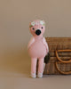 A hand knit pink Cuddle + Kind Penelope the Flamingo with a floral headband sits in front of a wicker basket on a beige background.