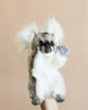 A hand is holding the Koala Puppet against a beige background. The fluffy, gray and white plush toy features large, fluffy ears, a dark nose, and small black eyes, giving it cute and realistic characteristics.