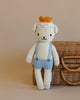 Sebastian the Lamb by cuddle+kind is a hand-knit, fair trade doll wearing light blue overalls and an orange crown, standing upright before a wicker suitcase on a beige background—a charming, sustainable creation.