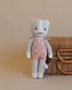 Cuddle + Kind Daisy the Kitten is a hand-knit gray cat doll made from premium cotton yarn, dressed in a pink dress with white flowers, standing upright before a wicker basket on a beige background.