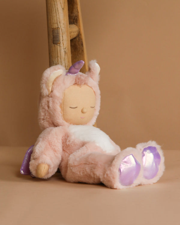 The Olli Ella Cozy Dinkum Doll Unicorn - Sugarpie features a soft pink body, horn, and shiny purple feet. With closed eyes, it exudes coziness as it leans against a wooden ladder on a warm beige background.