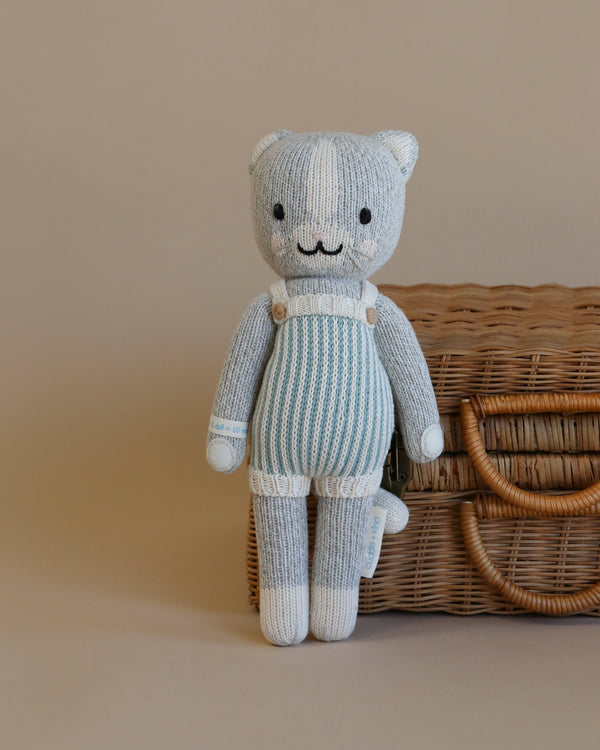 Cuddle + Kind Dylan the Kitten, a hand-knit gray cat plush in blue and white striped overalls, stands next to a closed wicker suitcase on a plain beige background.