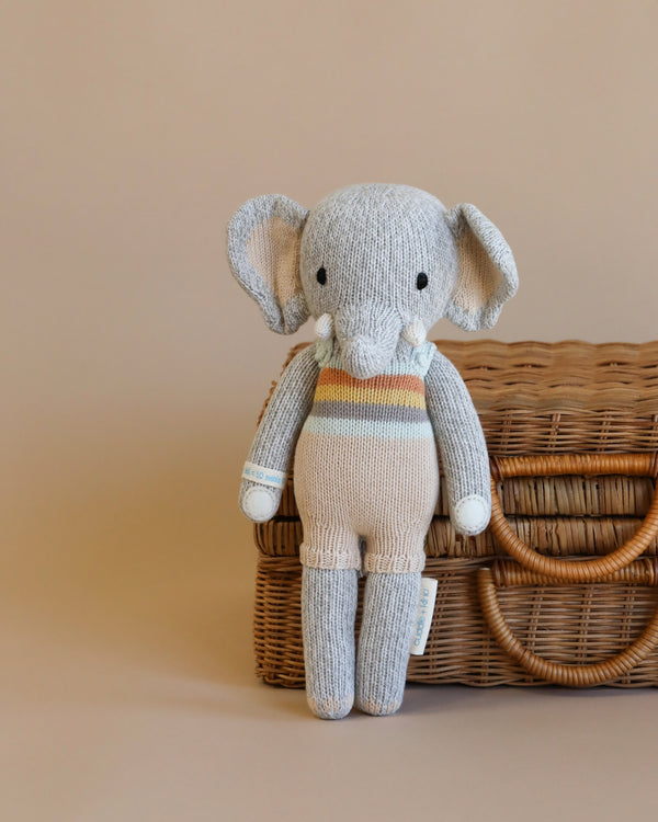 Cuddle + Kind Evan the Elephant is a hand-knit gray elephant doll with tusks and a striped outfit, shown standing in front of a woven wicker basket on a neutral beige background.