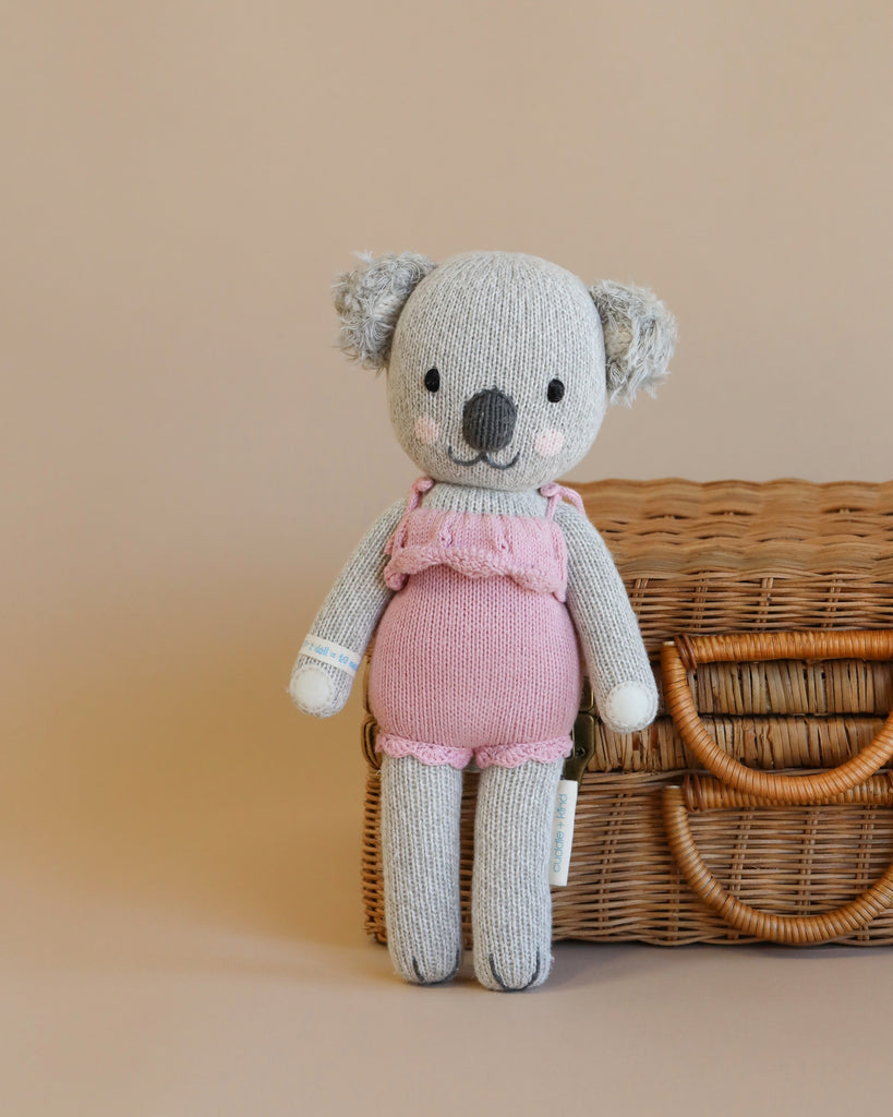 Cuddle + Kind Claire the Koala, a gray hand-knit doll in a pink outfit, is crafted with sustainable fair trade practices and pictured before a brown wicker basket on a neutral beige background.
