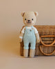 Cuddle + Kind Charlie The Honey Bear, a hand-knit teddy bear with a white shirt and light blue overalls, stands before a beige backdrop and woven wicker basket, filled with soft, hypoallergenic polyfill.