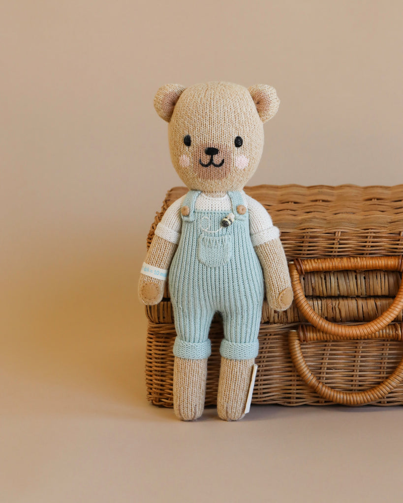 Cuddle + Kind Charlie The Honey Bear, a hand-knit teddy bear with a white shirt and light blue overalls, stands before a beige backdrop and woven wicker basket, filled with soft, hypoallergenic polyfill.