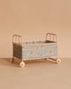 The Maileg Cot Bed, Micro - Powder features a light pink metal frame with wheels and blue floral fabric sides, and is shown on a beige surface against a matching beige background.